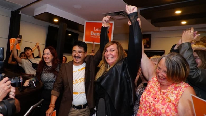 A woman smiles and pumps her fist in the air as others celebrate around her. Signs in the background say Leila Dance on them.