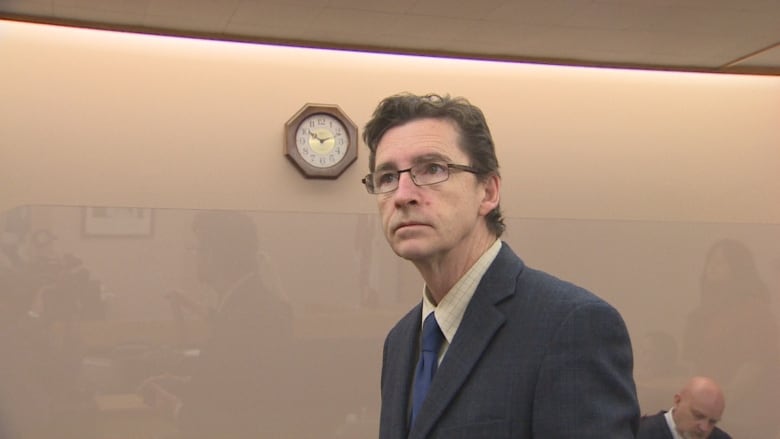 Man in grey suit and blue tie looks off in the distance of a beige coloured room. 