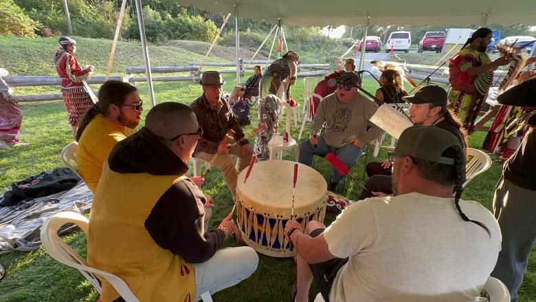 A group of men sit in a circle around a drum, each beating it with a stick and singing.