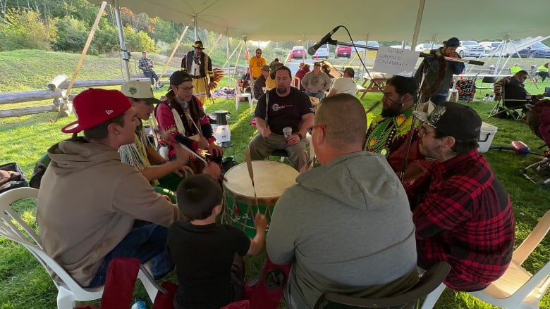 A group of men sit in a circle outside under a white tent beating a drum.