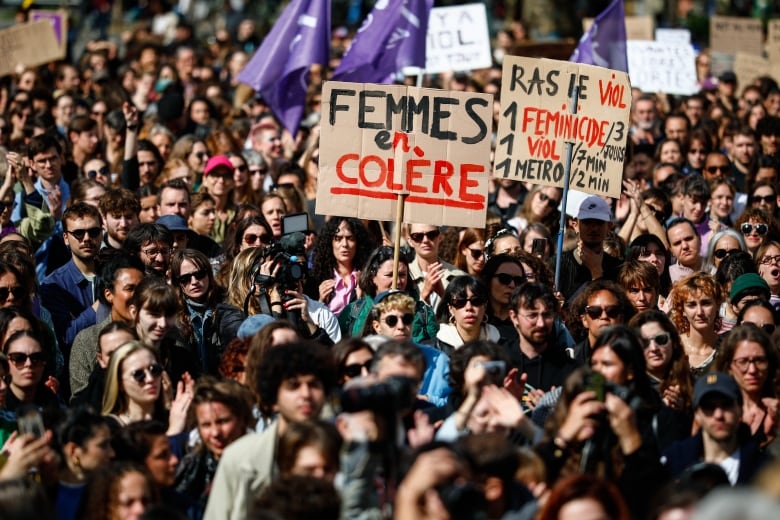 A huge crowd of people stand outside, some holding signs. One prominent sign, hand-painted on cardboard, reads: 