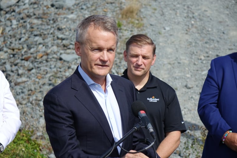 A man wearing a suit stands at a microphone in an off-road area.