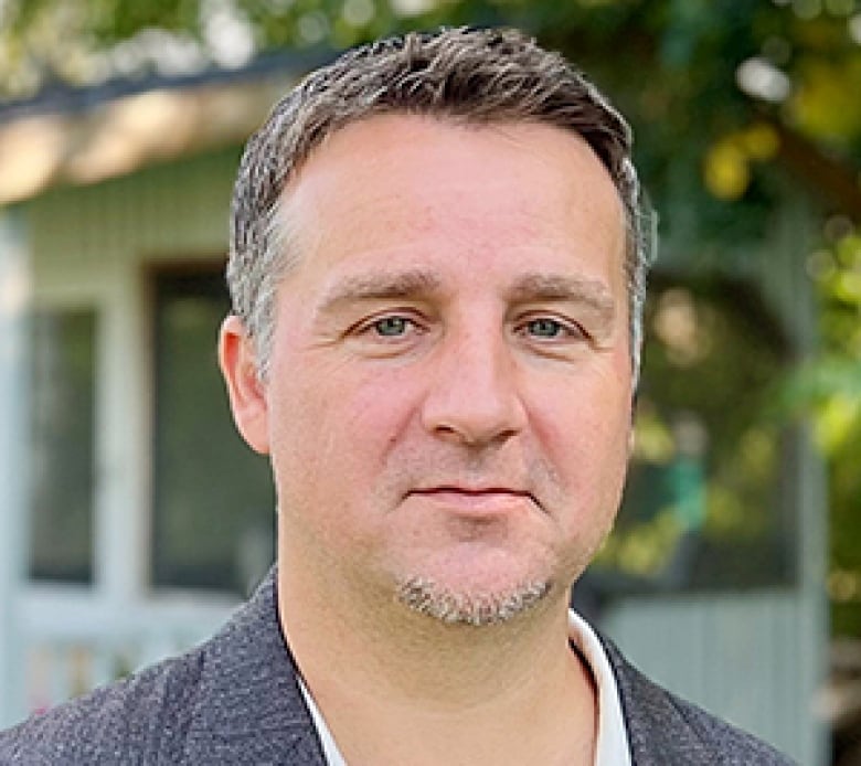 A man with short-cropped salt-and-pepper hair with a slight goatee, wearing a blue jacket and white shirt, looks at the camera.