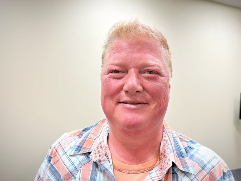 A woman with light blond hair and a red tanned face, wearing a plaid shirt, smiles.