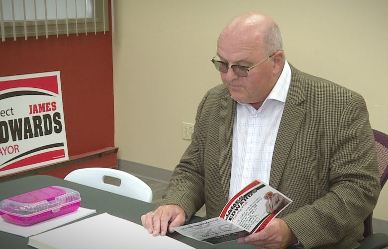 A man with an olive jacket and white shirt, wearing glasses, looks at an election brochure while sitting at a desk.