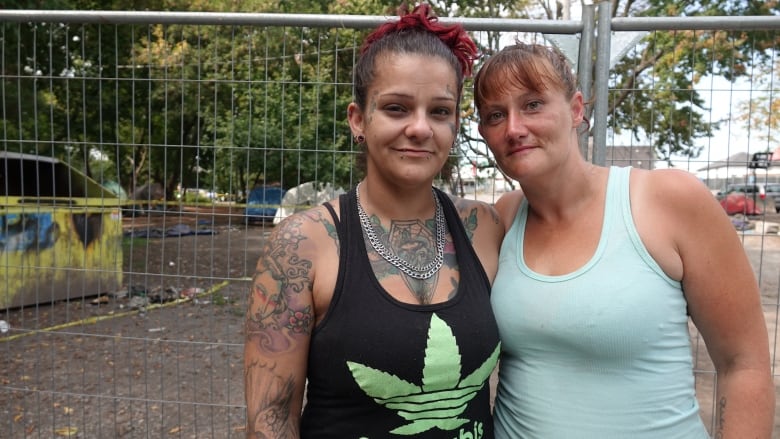 A woman with many tattoos and piercings hugs another woman with red hair and a blue tank top. A dumpster can be seen behind them and some tents.