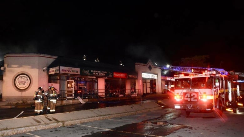 A night-time photo of a fire truck and firefighters standing outside a burnt strip mall.