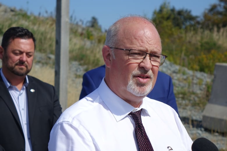 A man wearing a white shirt and black and red tie stands at a microphone on a sunny blue sky day.