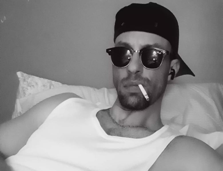 Man in sunglasses, ballcap on backwards and a cigarette hanging out of his mouth, reclining on a pillow. 