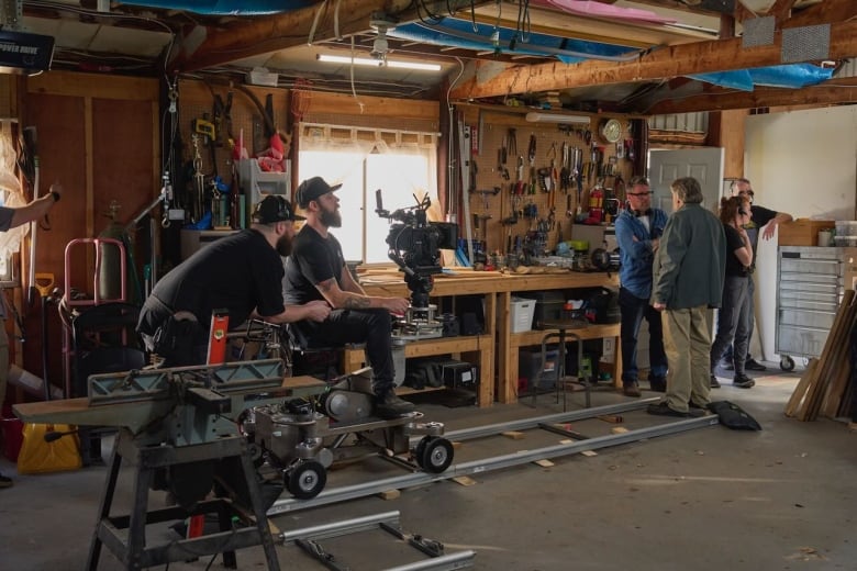 Two men in black t-shirts, beards and ballcaps sit in a room beside a movie camera on a dolly, on tracks in a room with wooden walls.