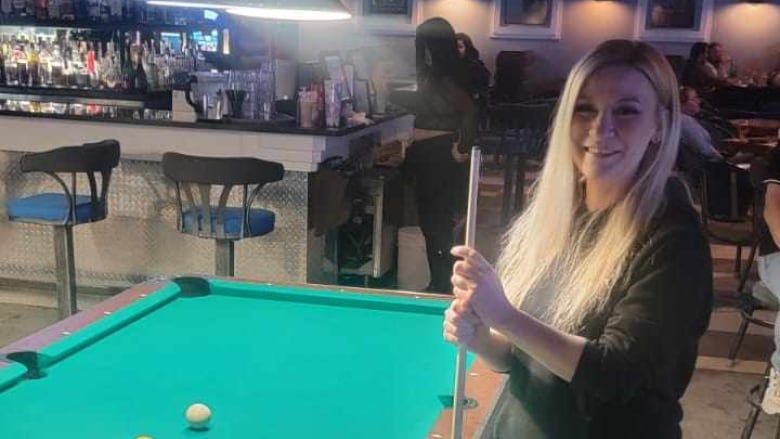 A woman stands beside a pool table holding a pool cue. 