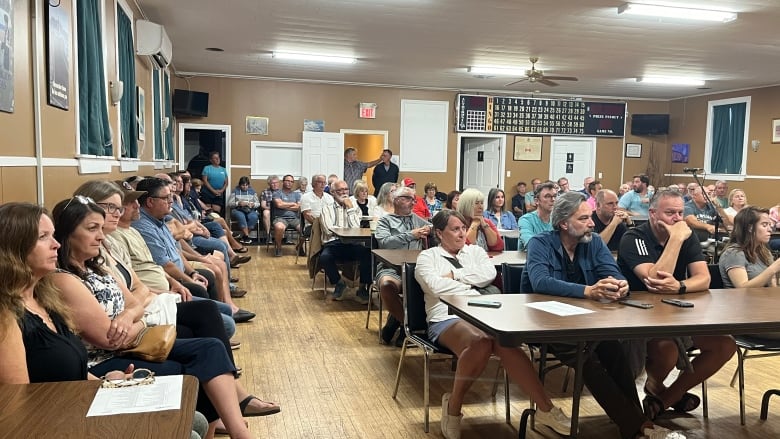 A large group of people sit behind tables and in chairs at a public meeting. 