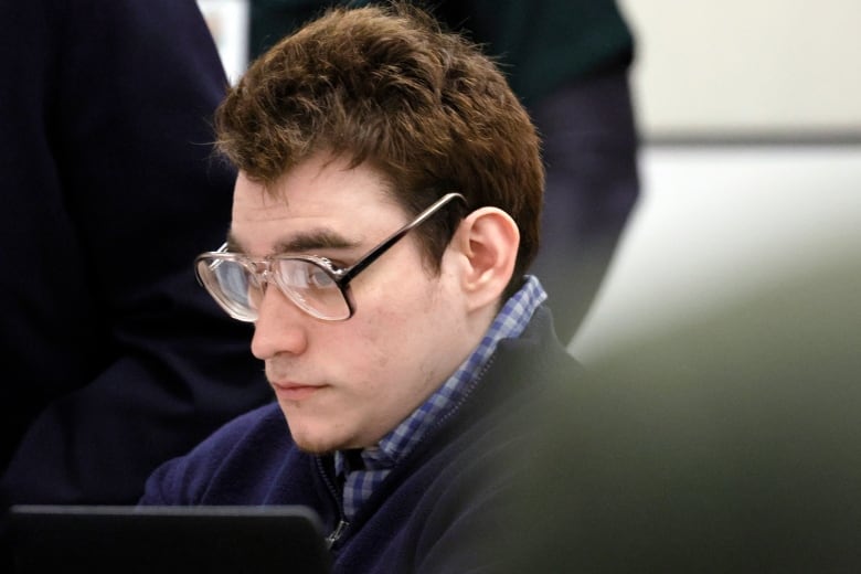 A young clean shaven, dark haired man with glasses is shown in closeup seated in a room.