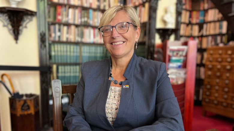 A woman wearing glasses and a suit smiles and places her hand one on top of another on a table, with rows of books in the background.