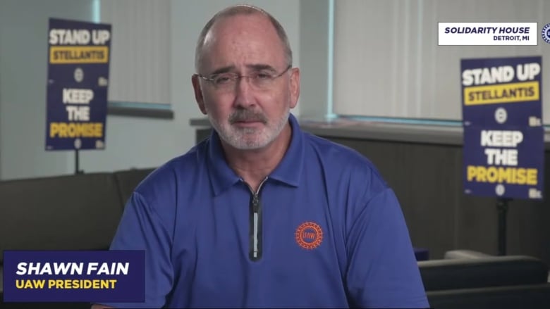 A man in a blue polo shirt sits in what appears to be an office looking at the camera while signs in the background read 