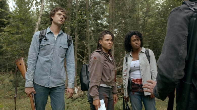 A man in a blue shirt holding a rifle, stands in the woods beside a woman in a brown shirt holding a map, and another woman in a white top and jacket.