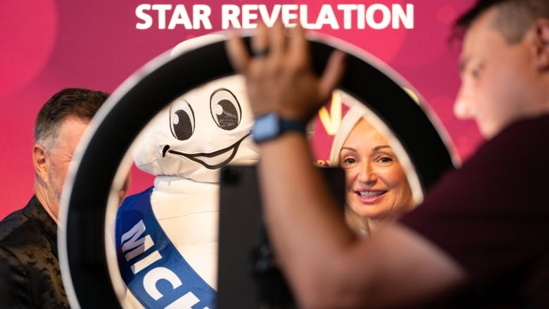 A blurry man in the foreground sets up a ring light in front of the Michelin Man, who is posing for a photo on the red carpet with a man and a woman