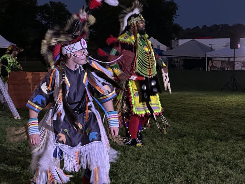 People dressed in colourful, traditional Indigenous clothing dance on grass at night time. 