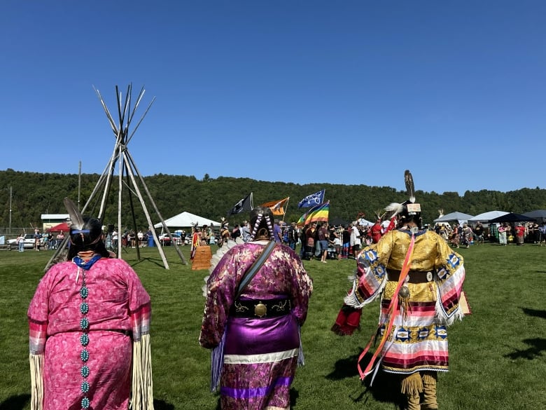 Three women dressed in colourful Indigenous regalia walk across a grassy lawn towards a crowd of people and a teepee.