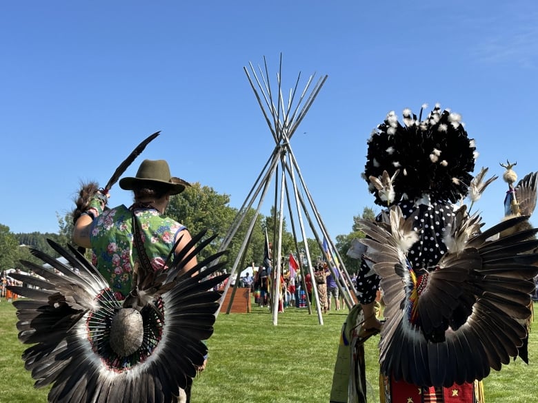 Two people dressed in traditional Indigenous regalia and feathers stand on a grassy lawn facing a wooden teepee.