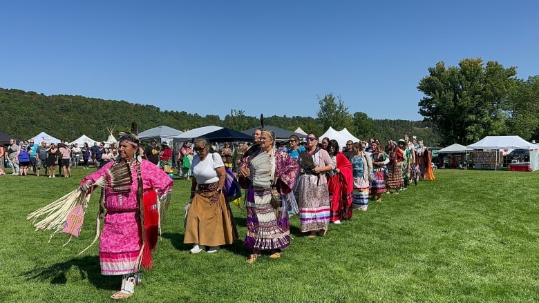 A group of people wearing Indigenous ribbon skirts walk in a line across a grassy lawn.