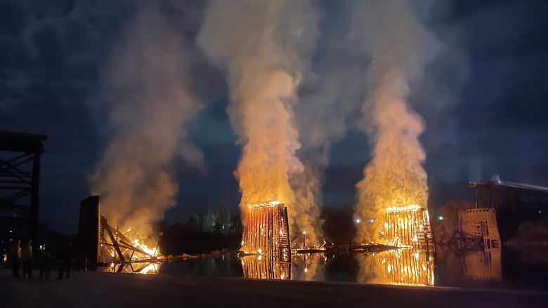 The remains of a wooden bridge in flames above a river with plumes of smoke rising into a dark sky.