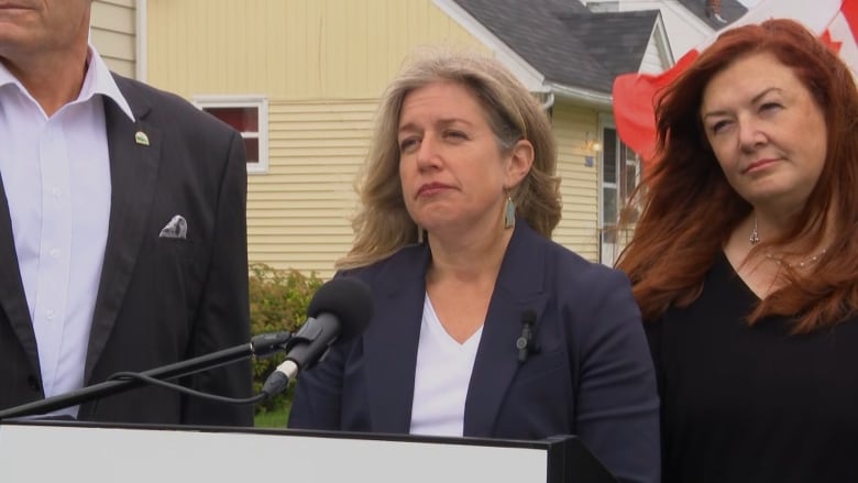 A blond woman in a blazer stands at a podium next to another woman with red hair.