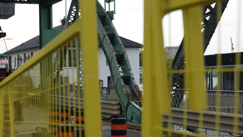 Two bent pieces of green metal are surrounded by other bridge elements as seen through a gap in a yellow security fence.