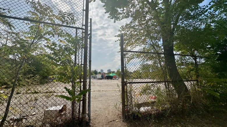 A metal gate open to a plot of land surrounded by vegetation with a tent and an RV sitting on the left area of it.