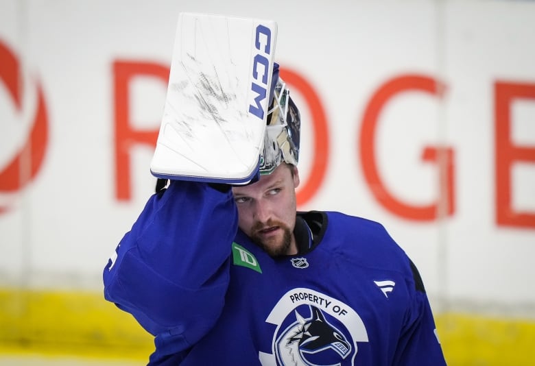 A goalie in blue training gear with the Canucks logo lifts his mask with a padded hand with a blurred-out Rogers logo on the white boards behind him.