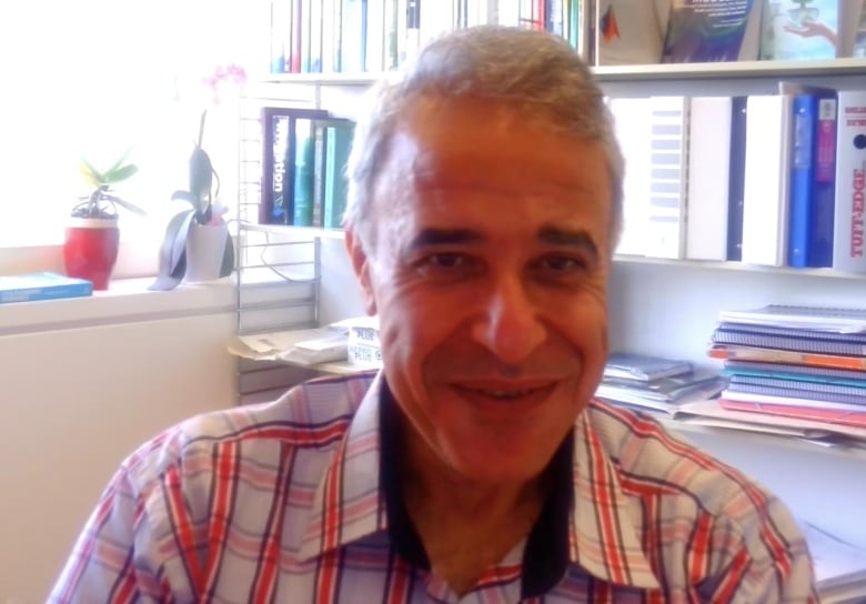 A man in a checkered red shirt with grey hair sits in front of a book shelf.