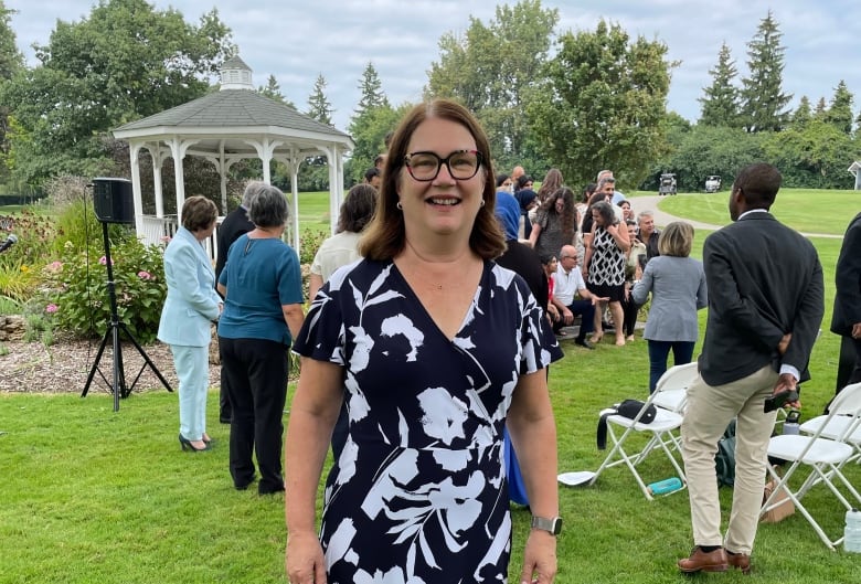 A woman in a black and white floral patterned dress poses on a lawn where other people, chairs and a pergola are seen in the background as an event unfolds.