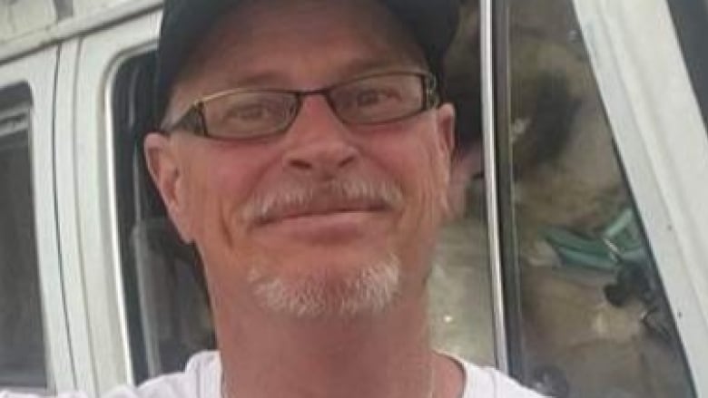 Close up of a white-haired man wearing a white shirt, a black baseball hat and glasses. 