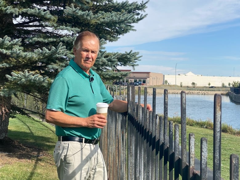 A man in a green shirt looks out at a lake, while holding a coffee cup.