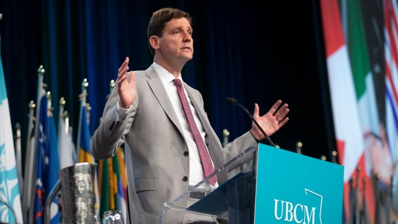 A tall man in a grey suit speaks on a stage. He gestures with his hands.