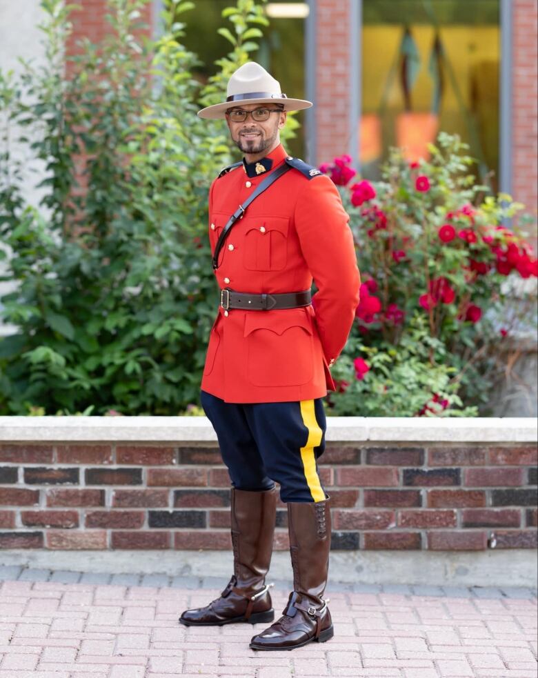 A man in RCMP ceremonial dress uniform stands on a brick sidewalk in front of some shrubs and a building.