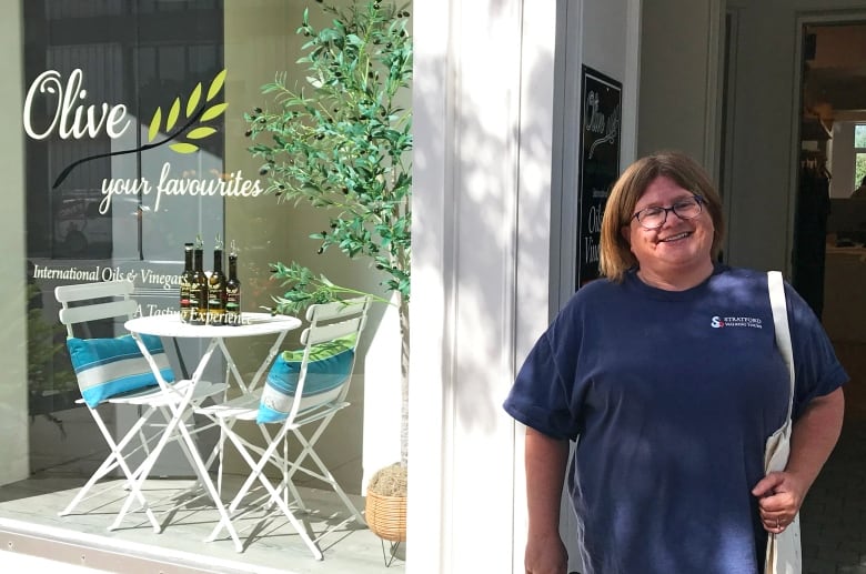 A woman wearing a blue shirt stands in front of an olive oil shop.