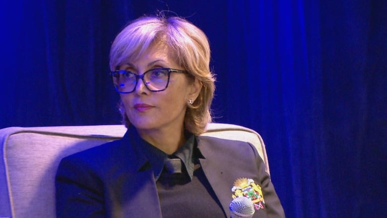 A woman with blond hair wearing black sits in front of a blue backdrop.