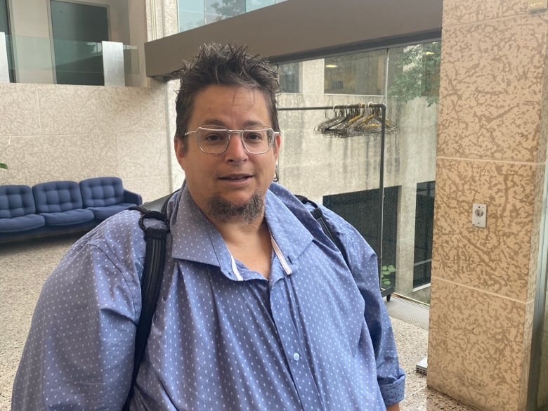 A person in a blue shirt and wearing glasses stands in the lobby of a building, backed by glass windows.