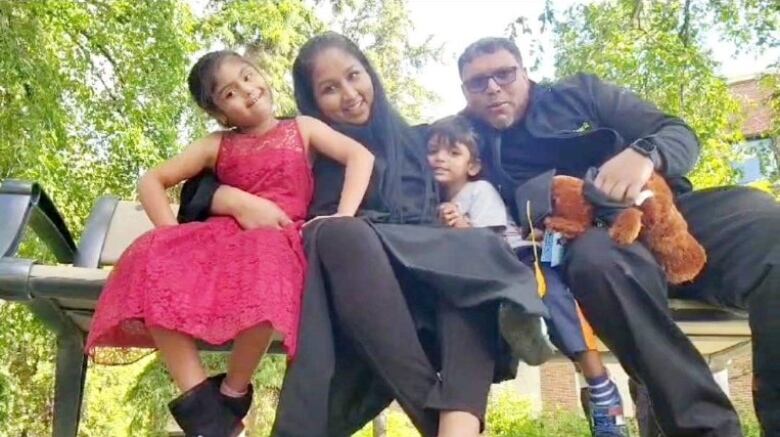 A husband and wife smiles on a park bench with their son and daughter. The husband holds a teddy bear wearing a graduation cap and the mother is wearing a graduation gown