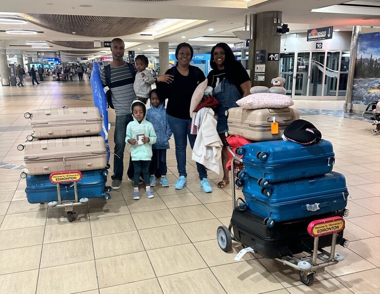 A mother and father pose with their three small children and two airport carts full of suitcases. A woman smiles next to them holding bags and smiling.