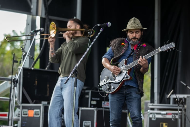 Two men performing on a stage, the one of the right is playing an electric guitar while the one on the right is playing an air instrument.