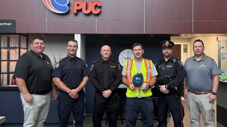 A man in a black shirt and tan pants stand at left, posing for a photo alongside a man in a black fire department uniform, another in a black paramedics uniform, another in a yellow safety vest, another in a black police uniform, and another in a blue shirt and tan pants.