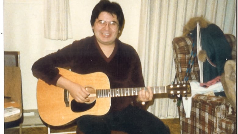 An old photo of a smiling Indigenous man sitting in an armchair holding an acoustic guitar.