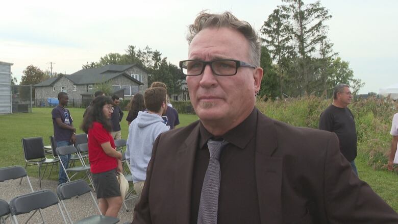 Head and shoulders shot of a man in a blazer standing outside with a lawn behind him. There are rows of empty folding chair and people standing up as if to leave.