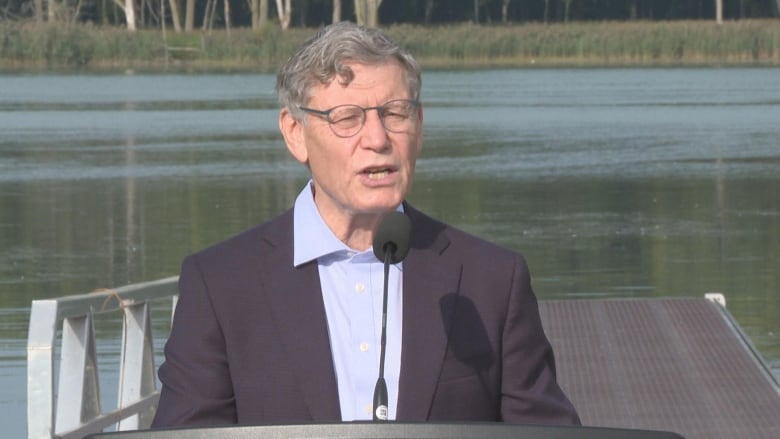 Head and shoulders shot of Terry at a podium with a lake behind him. 