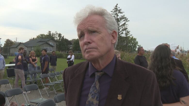 Close-up of Tim talking to reporters while people mill around behind him outdoors. 