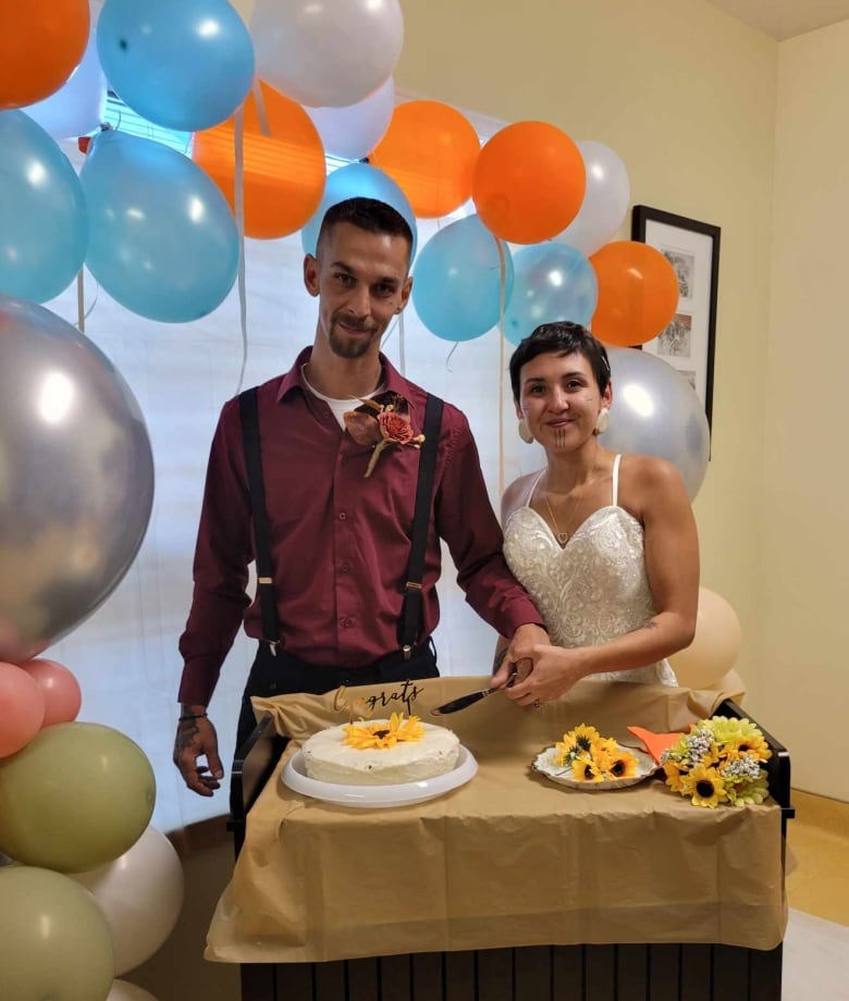 A woman in a wedding dress and man in maroon dress shirt cut a single layered wedding cake that has a single sunflower on top. There is a balloon arch behind them.