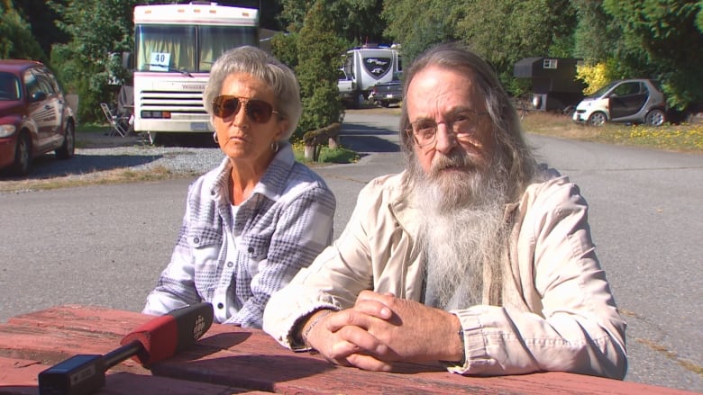 Two elderly individuals, one a man with a big white beard and another a woman in sun glasses sit down for an interview at an RV park.