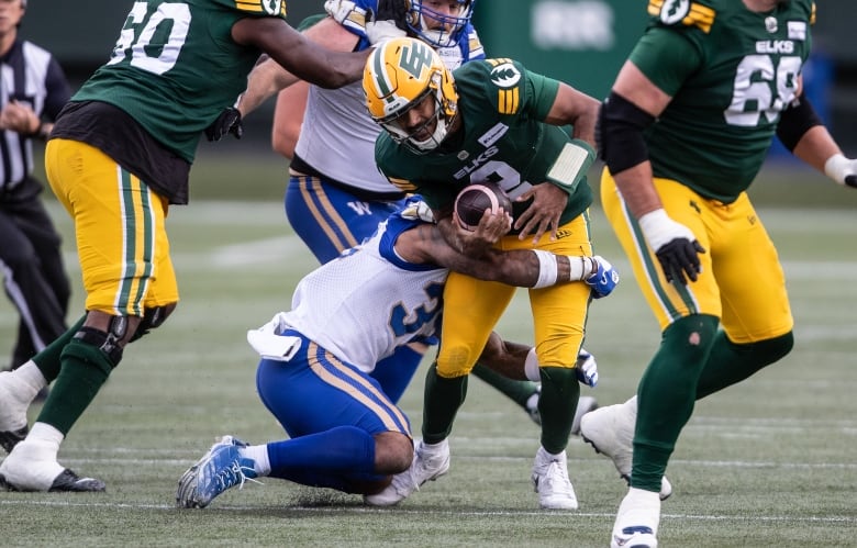 A football player in a blue and white uniform tackles a football player holding the football, wearing a green and yellow uniform. 